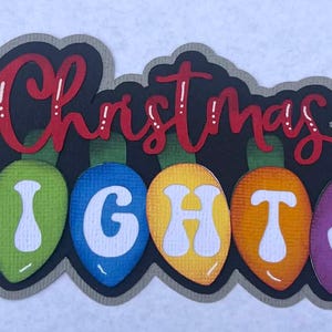 Könnte beinhalten: Eine farbenfrohe Weihnachtsdekoration mit den Worten "Christmas LIGHTS" in roter Schrift und weißen Blockbuchstaben. Die Buchstaben sind in einer Reihe von stilisierten, mehrfarbigen Glühbirnen angeordnet, mit einem schwarzen Hintergrund und einem grauen Rand.