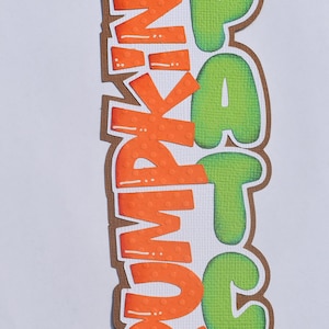 May include: A brown paper cutout of the words "Pumpkin Patch" with two orange pumpkins. The letters are in a 3D style and have a textured surface.