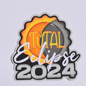 May include: A layered paper cutout design featuring the words "TOTAL Eclipse 2024". The design includes a stylized sun and moon in shades of orange, yellow, gray, and black, with a scalloped border.