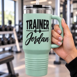 May include: A light mint green stainless steel tumbler with a handle and a clear lid. The tumbler features black text that reads TRAINER Jordan with a barbell icon between the two lines. The body has a ribbed texture at the bottom section.