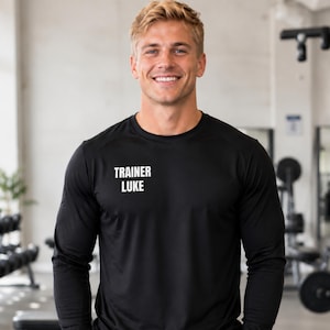 May include: A black long-sleeve athletic shirt featuring the white text TRAINER LUKE printed on the upper left chest area. The garment has a crew neckline and a smooth, moisture-wicking fabric construction suitable for active movement.