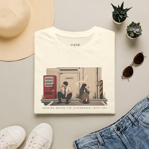 Op de afbeelding: Een crèmekleurig t-shirt met een grafische illustratie van twee personen die op de grond achter een gebouw zitten naast een rode automaat. De tekst "straykid" staat bij de kraag en "SMOKING BEHIND THE SUPERMARKET WITH YOU" staat onder de afbeelding gedrukt.