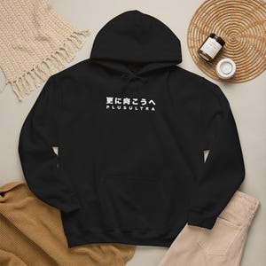 Peut inclure: Un sweat-shirt à capuche noir avec un texte blanc sur la poitrine indiquant "更に向こうへ" au-dessus du mot "PLUSULTRA". Le vêtement comprend une capuche à cordon, des manches longues et une poche kangourou, en tissu noir uni.