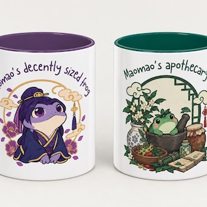 Könnte beinhalten: Zwei weiße Keramikbecher mit farbigen Rändern. Der linke Becher hat einen violetten Rand und eine violette Froschfigur in traditioneller Kleidung mit dem Text Maomao's decently sized frog. Der rechte Becher hat einen grünen Rand und einen Frosch.