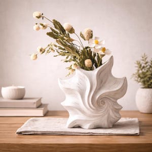 May include: A white, sculptural vase with a swirling design, holding dried flowers and greenery. The vase sits on a neutral-colored linen placemat on a wooden table. A small candle and books are in the background.