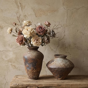 Puede incluir: Dos jarrones de cerámica rústica, uno alto y otro bajo, sobre una superficie de madera envejecida. El jarrón alto contiene un ramo de flores artificiales en crema, malva y gris. El fondo es una pared texturizada de tonos neutros.