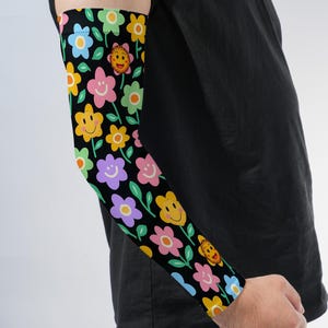 May include: An arm sleeve featuring a black background with a repeating pattern of colorful flowers. Each flower has a simple smiling face with two eyes and a curved mouth. The flowers are rendered in shades of yellow, pink, light blue, and light green.