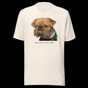 May include: A cream-colored short-sleeve t-shirt featuring a photographic print of a brown Brussels Griffon dog wearing a black harness. Below the dog, the text "this was not my idea" is printed in a small, simple, black, lowercase font.