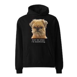 May include: A black hooded sweatshirt featuring a graphic of a tan, scruffy-faced dog. Below the dog, the text reads: leave me alone. i'm manifesting. The hoodie includes long sleeves, a front kangaroo pocket, and a drawstring hood.