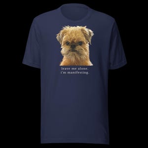 May include: A navy blue short-sleeved t-shirt featuring a photographic print of a tan, scruffy-haired dog. Below the dog, the text reads: leave me alone. i'm manifesting. The shirt has a crew neckline and a standard fit, showcasing a clear graphic.