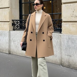 May include: A tan double-breasted coat with black buttons. The coat is worn over a cream-coloured turtleneck and light green trousers. A black handbag hangs from the shoulder. The person is wearing black sunglasses.