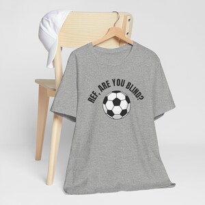 May include: A heather gray short-sleeve t-shirt featuring a black graphic of a soccer ball centered on the chest. Above the soccer ball, the text reads "REF, ARE YOU BLIND?" in a bold, black, sans-serif font. The shirt is displayed on a wooden hanger.