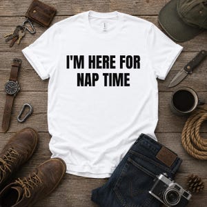 Könnte beinhalten: Ein weißes kurzärmeliges T-Shirt mit dem schwarzen Text I'M HERE FOR NAP TIME, der mittig aufgedruckt ist. Das Shirt verfügt über einen Rundhalsausschnitt und eine Standardpassform und wird flach liegend mit klarem Design präsentiert.