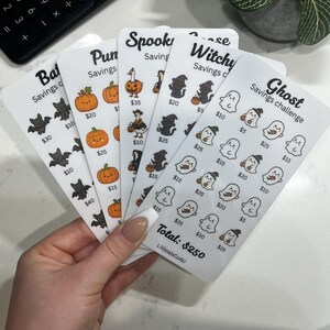 May include: A set of five white paper savings challenge trackers featuring Halloween-themed illustrations including bats, pumpkins, geese, cats, and ghosts. Each tracker displays specific dollar amounts under the icons and the text LilGoalsCoAU.