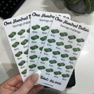 May include: Three white rectangular savings challenge cards featuring the text One Hundred Dollar Savings challenge and a grid of green dollar bill icons. Each icon is labelled with $100. The bottom of the card displays Total: $2,000 and LilGoalsCoAU.