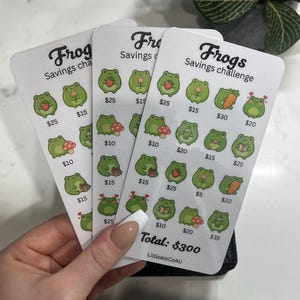 May include: Three white rectangular savings challenge cards featuring a "Frogs Savings challenge" header and multiple small green frog illustrations. Each frog is paired with a dollar amount ranging from $5 to $35. The bottom text reads "Total: $300" and "LilGoalsCoAU".