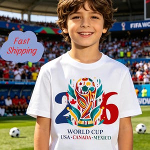 May include: A white short-sleeved t-shirt featuring a colorful graphic of the World Cup trophy centered between the numbers 2 and 6. Below the trophy, the text reads WORLD CUP USA CANADA MEXICO in blue, red, and green lettering.