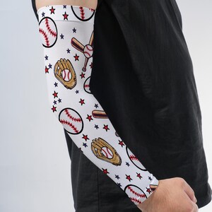 May include: A white compression arm sleeve featuring a repeating pattern of baseballs, brown baseball gloves, wooden bats, and small red and blue stars. The sleeve is designed to cover the forearm and bicep area with a snug, athletic fit.