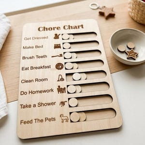 May include: A light-coloured wooden chore chart featuring the text "Chore Chart" at the top. It includes a list of tasks such as "Get Dressed," "Make Bed," "Brush Teeth," "Eat Breakfast," "Clean Room," "Do Homework," "Take a Shower," and "Feed The Pets," each paired with a small icon and a horizontal sliding track with a circular wooden slider.