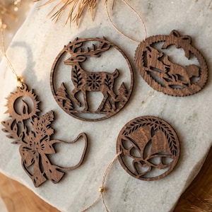 May include: Four dark brown laser-cut wooden ornaments featuring intricate holiday designs. One is a circular deer motif, another a sleigh, one a candle with pine branches, and the last a circular bell design. Each includes a thin gold hanging string.