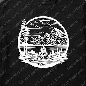May include: Black t-shirt featuring a white circular graphic of a mountain landscape with a campfire, lake, and trees. The text "Larsen Made Crafts" is printed around the shirt's collar.