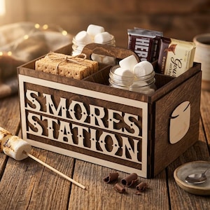 Puede incluir: Una caja de madera de S'mores Station con compartimentos para galletas Graham, malvaviscos y barras de chocolate. La caja tiene las palabras "S'MORES STATION" recortadas en la parte delantera. También se ve un malvavisco asado en un palito y una taza de café.