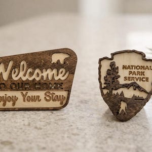 May include: Two wooden signs with a rustic finish. One sign reads "Welcome to our home, Enjoy Your Stay" with a bear silhouette. The other sign is a National Park Service emblem.