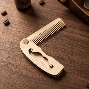 May include: A wooden folding comb with a mustache cutout, displayed open on a wooden surface. A small bottle of beard oil and coffee beans are also visible. The comb is light brown and has fine teeth.