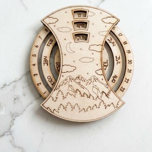 May include: A circular wooden perpetual calendar featuring laser-engraved mountain and forest scenery. The design includes rotating rings for dates, months, and days of the week, with windows displaying 12, APR, and SUN. The light wood surface is etched with clouds, stars, and a crescent moon.