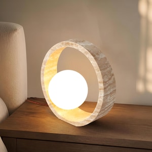 May include: A circular table lamp featuring a natural beige stone frame with a textured, marbled appearance. A smooth, spherical white glass bulb is positioned in the center of the ring, emitting a warm, soft glow that illuminates the surrounding area.