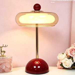 Könnte beinhalten: Eine Tischleuchte mit einem glänzend roten Kuppelsockel, einem schlanken goldfarbenen Metallschaft und einem breiten, transparenten bernsteinfarbenen Glasschirm mit einem cremefarbenen inneren Lichtdiffusor. Ein kleiner roter Zierknopf sitzt oben.