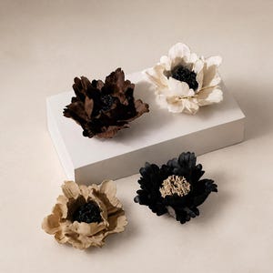 May include: Four decorative flower ornaments featuring layered petals. Two flowers are dark brown and black with dark centers, one is cream with a black center, and one is light tan with a dark center. The flowers are arranged on and around a white block.