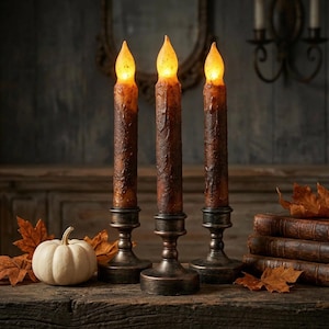 Peut inclure: Trois bougies effilées à piles avec une finition en cire brun foncé texturée et des pointes en forme de flamme orange lumineuse. Chaque bougie est placée dans un support sur pied en métal foncé, disposées en rangée avec une petite citrouille.