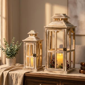 May include: Two wooden lanterns with a distressed white and brown finish. The lanterns feature glass panels, hinged doors with small metal latches, and tiered roofs. Each lantern contains a pillar candle, with the larger one including greenery.