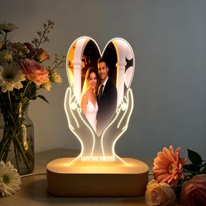 Op de afbeelding: Een hartvormige LED-lamp met een foto van een stel, vastgehouden door verlichte handen. De basis is van hout en toont de tekst "LOVE YOU FOREVER". De lamp wordt omringd door bloemen, wat een romantische sfeer creëert.