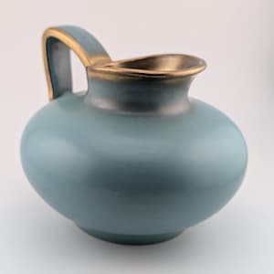 Op de afbeelding: Een kleine, ronde keramische kan met een matte, stoffig blauwe afwerking. Het object heeft een gebogen handvat en een brede, open tuit, beide geaccentueerd met een metallic gouden rand langs de randen, wat een contrast vormt met het blauwe lichaam.