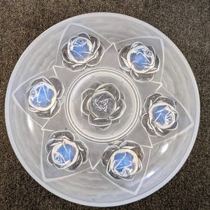 May include: A circular clear glass serving plate featuring a frosted finish and a central embossed design of seven roses. Six roses are arranged in a hexagonal pattern around a single larger rose in the centre, all set against a star-shaped base.