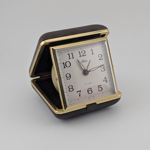 May include: A portable travel alarm clock with a dark brown leather-like folding case and a gold-tone metal frame. The square clock face features a white background, black Arabic numerals, the Sears logo, and black hour and minute hands.