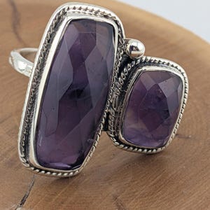 Puede incluir: Un anillo de plata con dos piedras de amatista rectangulares y facetadas. La piedra más grande está orientada verticalmente, la más pequeña a la derecha. Ambas están engastadas en un bisel detallado en forma de cuerda.