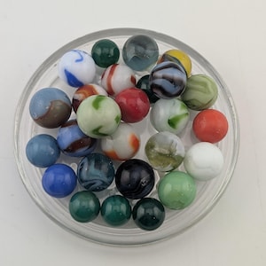 May include: A collection of assorted glass marbles in a clear circular glass dish. The marbles feature various colours including blue, green, red, white, black, and grey, with swirling patterns, solid opaque finishes, and translucent glass textures.