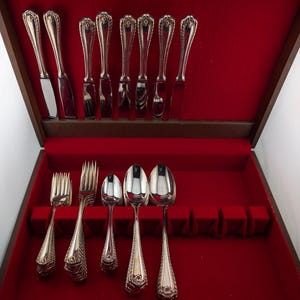 May include: A set of silver-toned cutlery arranged in a wooden storage box with a red velvet interior. The collection includes eight knives with decorative handles in the top row, and two forks and three spoons in the bottom row.