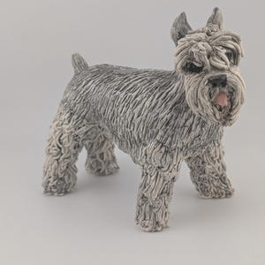 May include: A ceramic Schnauzer dog sculpture in grey and white. The dog has a textured coat, a pink nose, and a distinctive beard. The sculpture is standing on all fours, with its tail slightly raised. The sculpture is a decorative item.