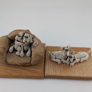 May include: Two ceramic dog figurines on a wooden base, one set featuring two dogs sitting together in a textured armchair, and the other set featuring two dogs lying down on a flat wooden square. The dogs are light gray with dark noses.