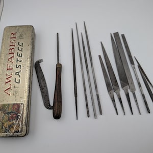 Puede incluir: Una caja de almacenamiento rectangular de metal A.W. FABER CASTELL con una ilustración vintage, acompañada de una colección de varias limas de metal y una herramienta con mango de madera y hoja de metal curva, dispuestas sobre una superficie lisa.