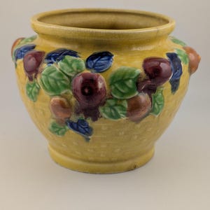 Peut inclure: Un cache-pot en céramique jaune avec un motif extérieur en vannerie texturée. Le récipient présente des décorations en relief tridimensionnelles représentant des baies rouge foncé, des fruits bruns et des feuilles vertes sur le pourtour.