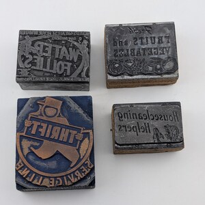May include: Four rectangular metal printing blocks with wooden bases. One block features a figure in a top hat with the text THRIFT and REBAICE. Another shows text reading LOLLIES. A third displays FRUITS AND VEGETABLES. The fourth shows Housecleaning Helpers.