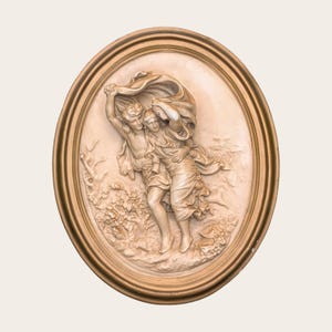 Puede incluir: Una placa de pared ovalada con un relieve de color crema de dos figuras envueltas en telas fluidas, encerradas en un marco de color dorado metálico. Las figuras se muestran en movimiento, con la tela ondeando sobre sus cabezas.