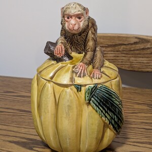 May include: A ceramic jar shaped like a yellow pumpkin with a lid featuring a sculpted brown monkey sitting on top. The pumpkin has vertical ridges and a single green leaf attached to the side. The monkey has light brown fur and pinkish skin.