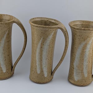 May include: Three tall, cylindrical ceramic mugs with a speckled tan finish and a single curved handle. Each mug features two vertical, white, brush-stroke style markings on the front. The mugs have a flared rim and a smooth, tapered body.