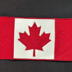 May include: Embroidered rectangular patch featuring the Canadian flag. The flag has a red background with a white square in the center, displaying a red maple leaf. The patch is likely for clothing or accessories.